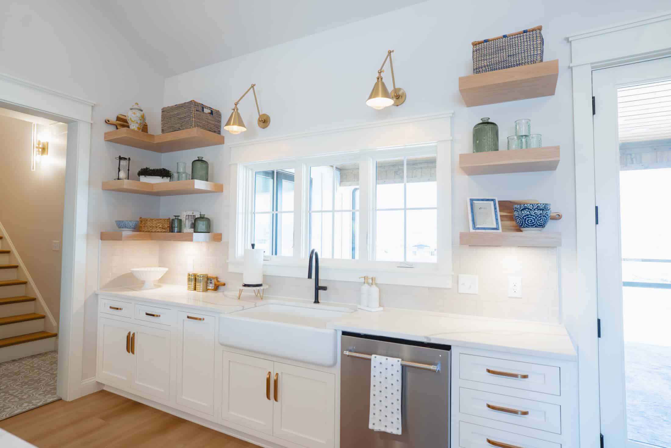 White modern farmhouse kitchen with apron-front sink, matte black faucet, floating wood shelves and brass wall sconces.