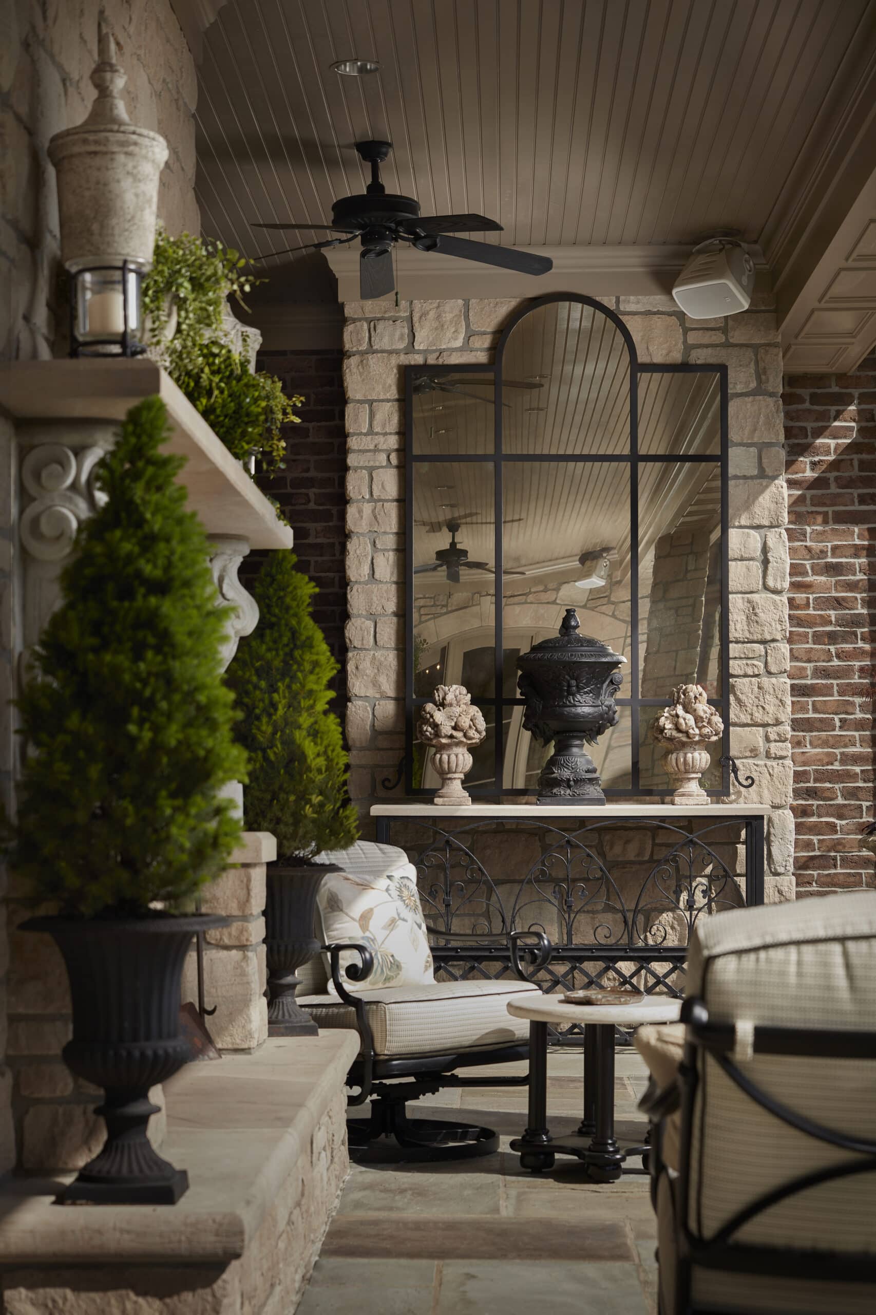 Covered stone-walled patio with large arched mirror, black ceiling fan, wrought-iron table, cushioned chairs and potted topiaries.