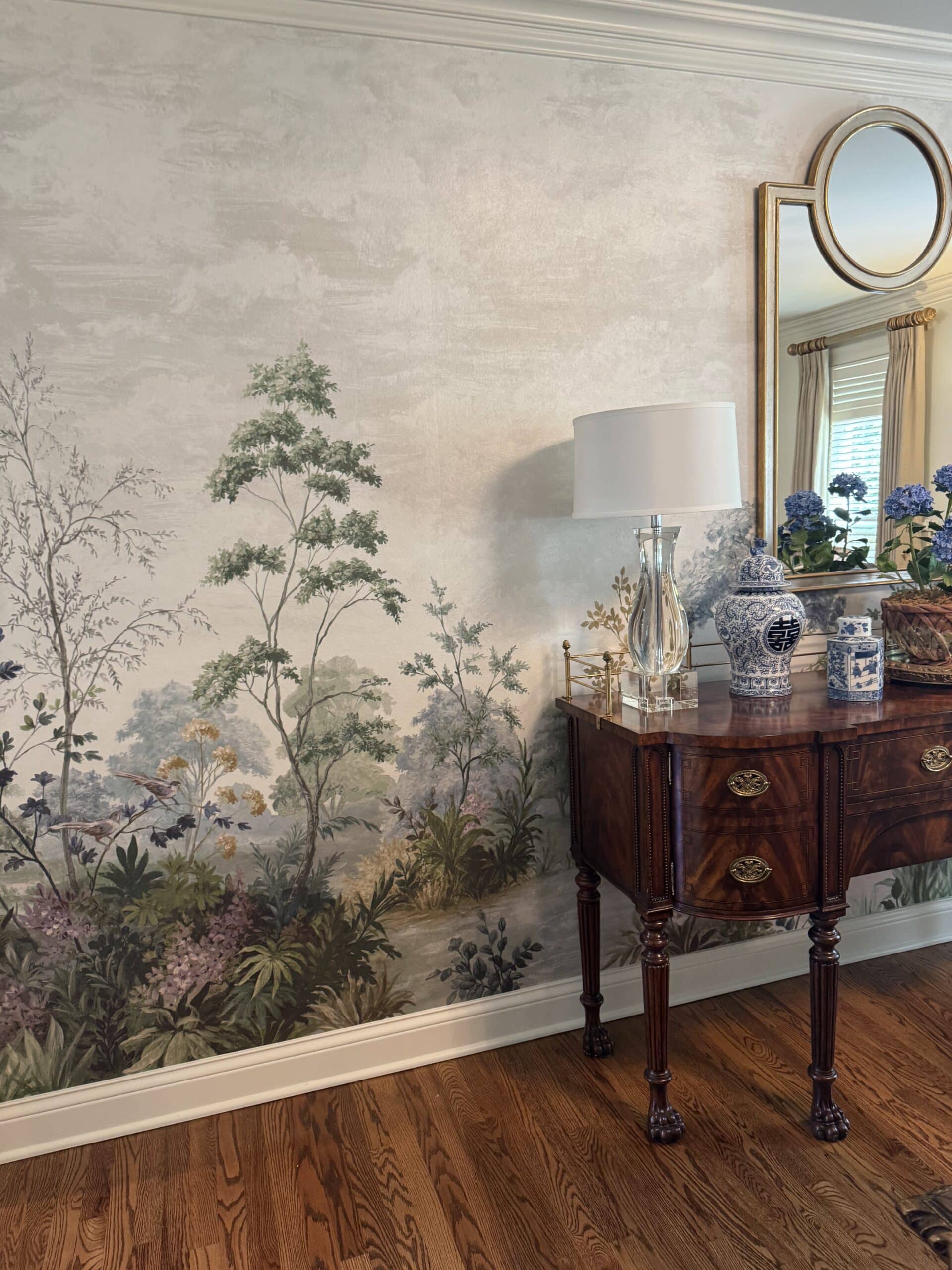 Botanical wallpaper mural with painted trees, antique mahogany side table, glass lamp and blue-and-white ginger jar.