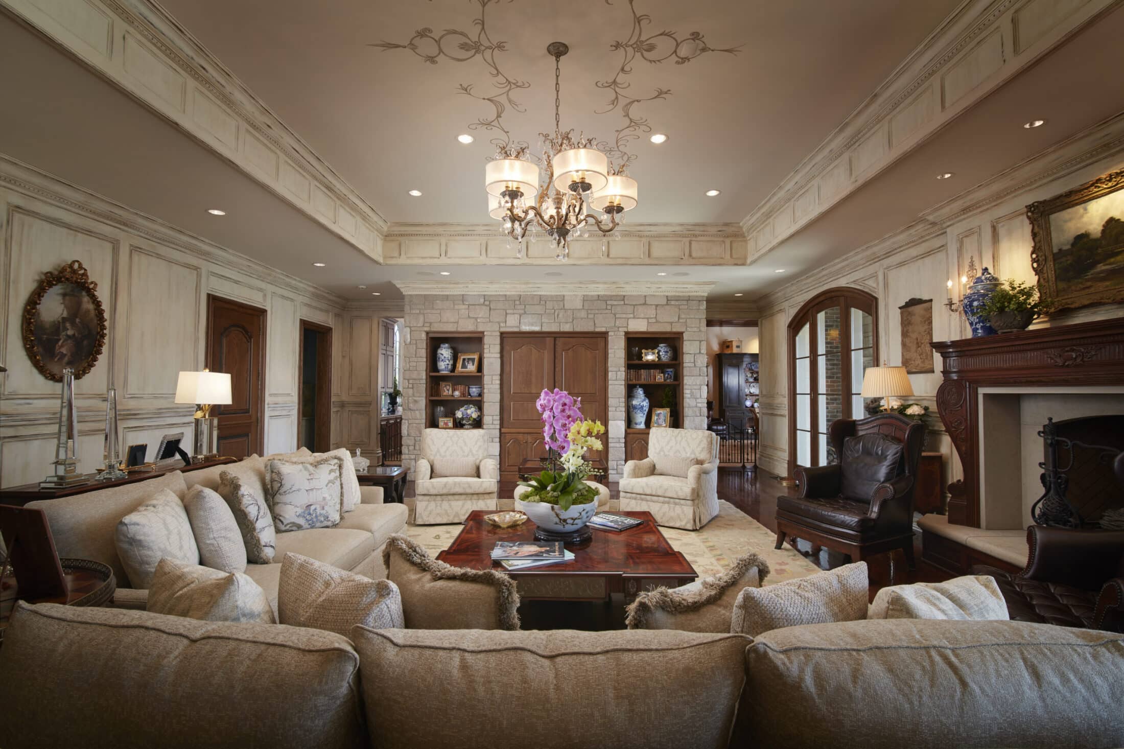 Luxury traditional living room interior with beige seating area, crystal chandelier, wood coffee table, pink orchids and stone accent wall.